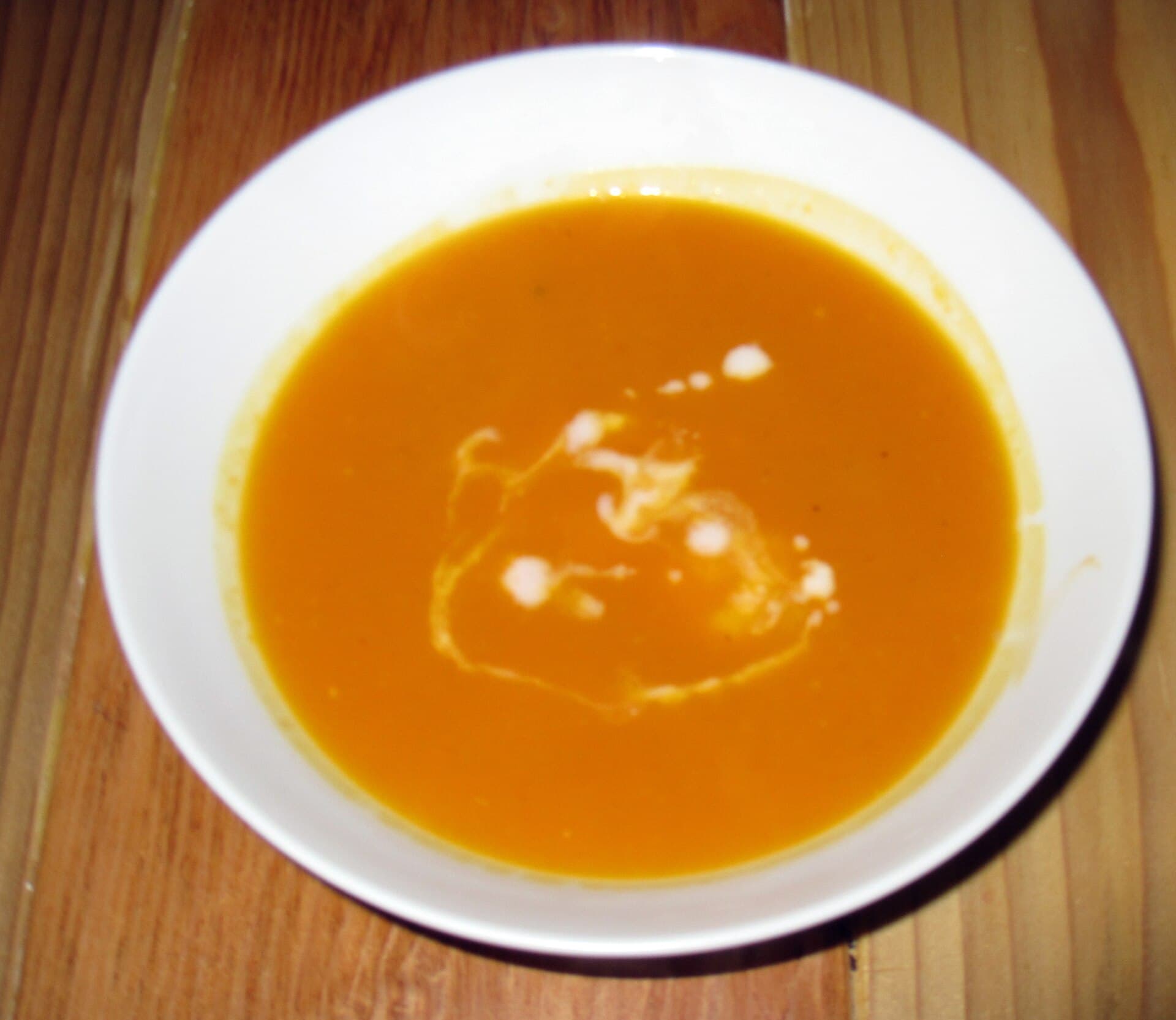 A white bowl of smooth orange butternut squash soup with a swirl of cream, set on a wooden table.